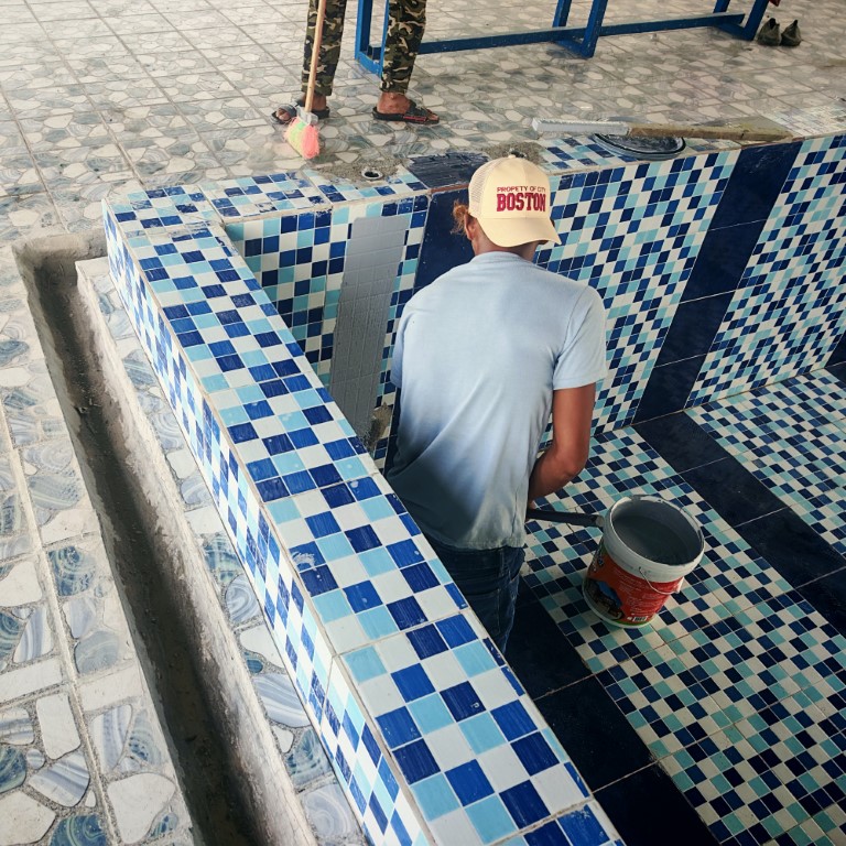 A pool repair specialist carefully applying gray waterproof grout to blue checkered mosaic tiles during a swimming pool leak repair.