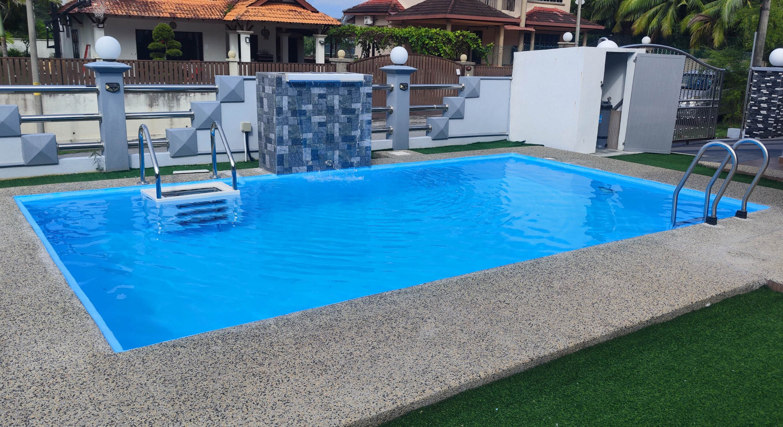 A bright blue rectangular fiberglass swimming pool surrounded by pebble decking and artificial grass. It features a grey stone waterfall, built-in white entry steps, and stainless steel handrails in a residential yard.