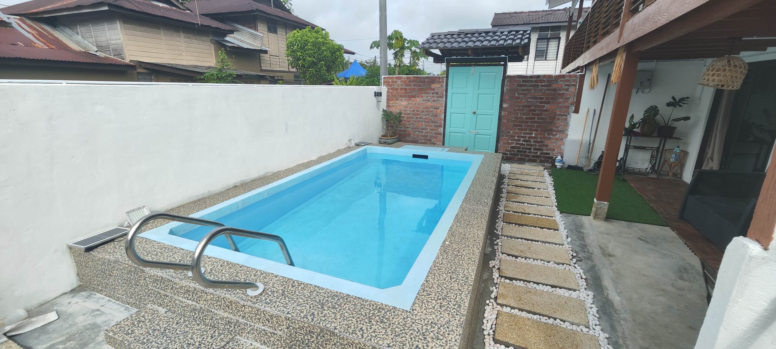 A light blue rectangular fiberglass pool with a raised pebble edge. It sits between a white boundary wall and a traditional wooden house, featuring stepping stones and white garden pebbles.