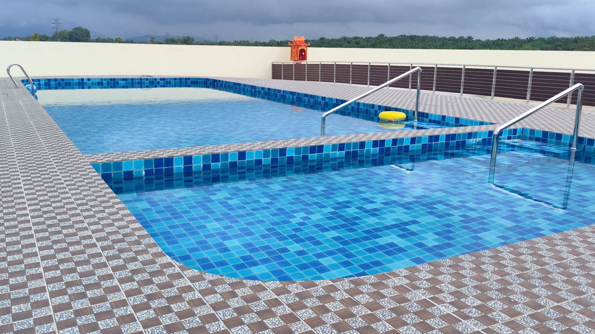 A custom dual-zone concrete pool with blue mosaic tiles and stainless steel handrails, set into a checkered tile deck overlooking a green tree line.